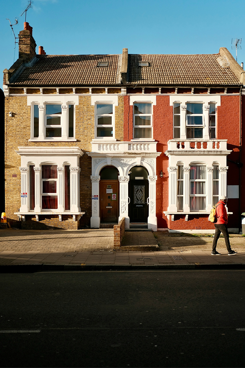 A pair of Victorian semi-detached houses, one in yellow brick and one painted red, with white bay windows and ornate arched doorways, as a person in a red jacket walks past on the pavement.