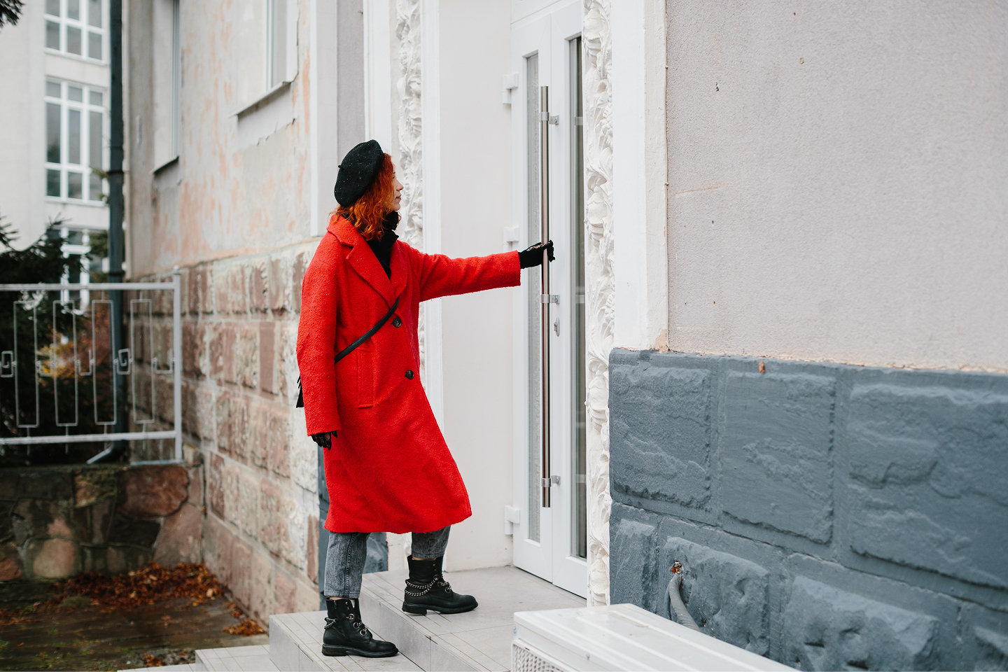 A woman with red hair, wearing a long red coat, black beret, jeans and black boots, stands on a doorstep reaching for the handle of a white front door with ornate stone surrounds.