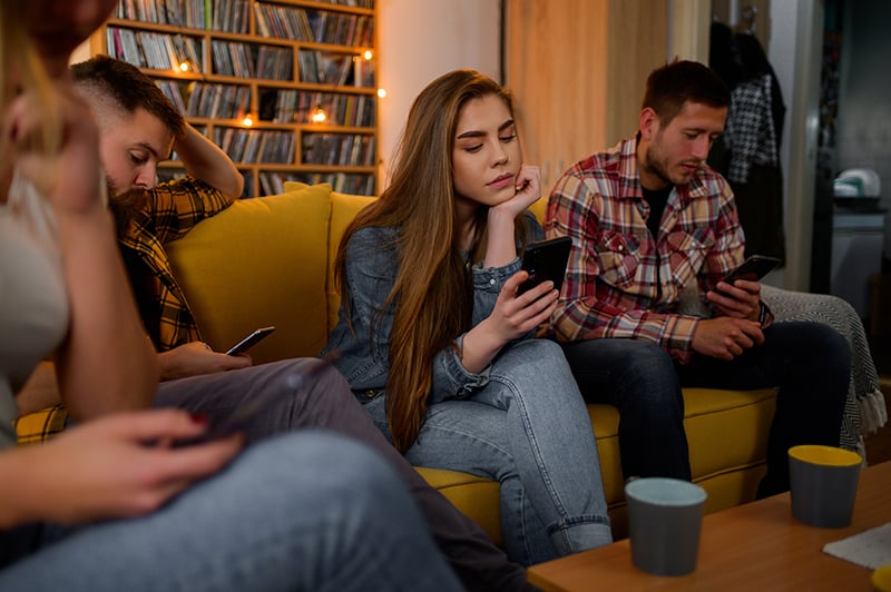 A group of young adults sitting on a yellow sofa in a cosy living room, each looking at their own phone, with a bookshelf and warm fairy lights behind them.