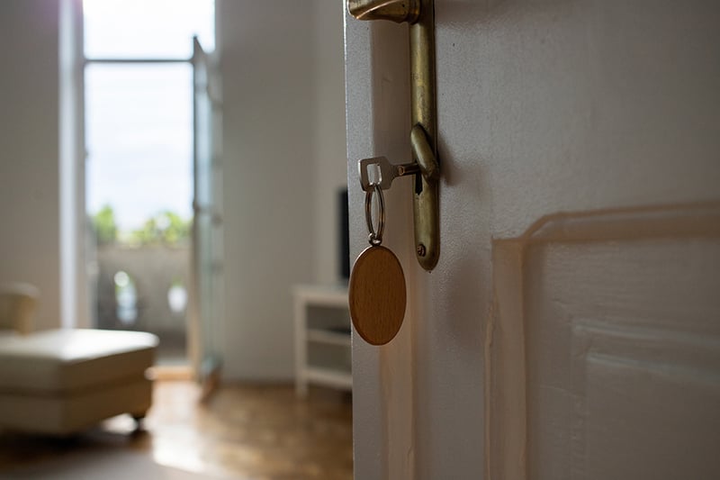 A key with a wooden fob hanging in the brass lock of an open white door, with a bright, sparsely furnished room and window blurred in the background.