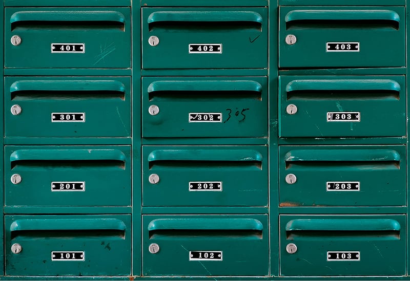 A wall of worn green metal mailboxes in a grid, each with a lock and a numbered flat label from 101 to 403, one with “305” handwritten beside its label.