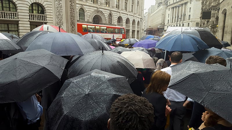 A dense crowd of people holding rain-spotted umbrellas, mostly black, on a wet city street, with a red double-decker bus and grand stone buildings in the background.
