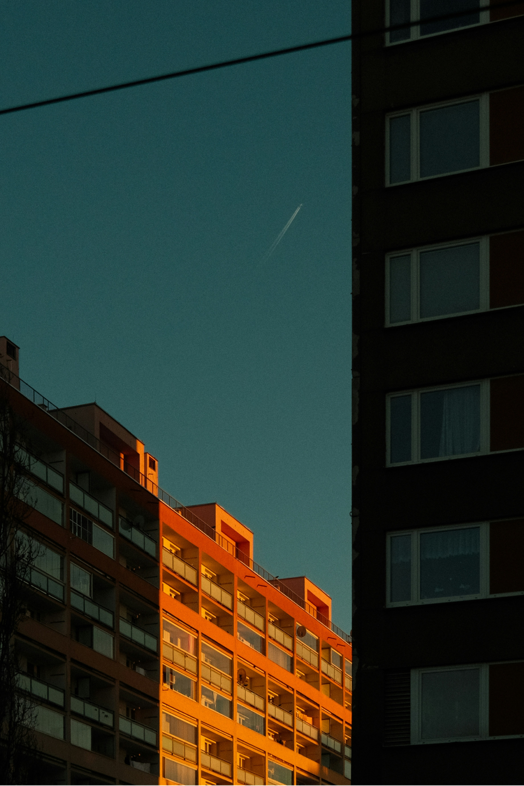 A block of flats with rows of balconies lit orange by low sunlight, beside a darker residential tower in shadow, under a deep teal sky with an aeroplane contrail.