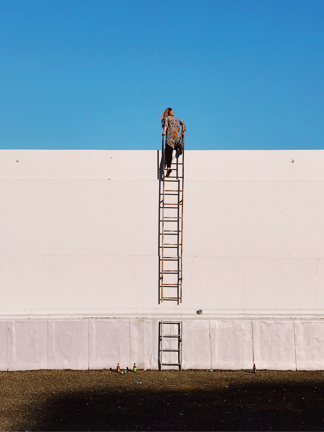 A person climbing a tall metal ladder up the side of a large white wall, nearly at the top, against a clear blue sky.