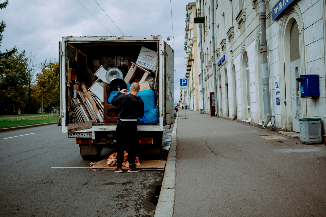 A man loading or unloading a removal van packed with boxes, furniture and wrapped belongings, parked at the kerb of a quiet city street lined with tall buildings.