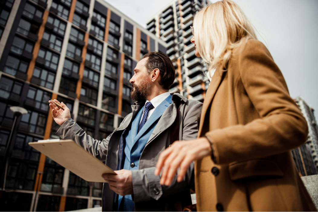 A bearded man in a suit and grey coat, holding a clipboard, gestures towards a modern high-rise apartment block while talking with a blonde woman in a camel coat.