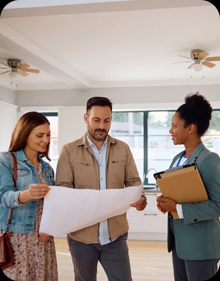 A real estate agent, a woman, talks with a couple, a man and a woman, who are reviewing a large blueprint or floor plan in an empty, bright room of a house.