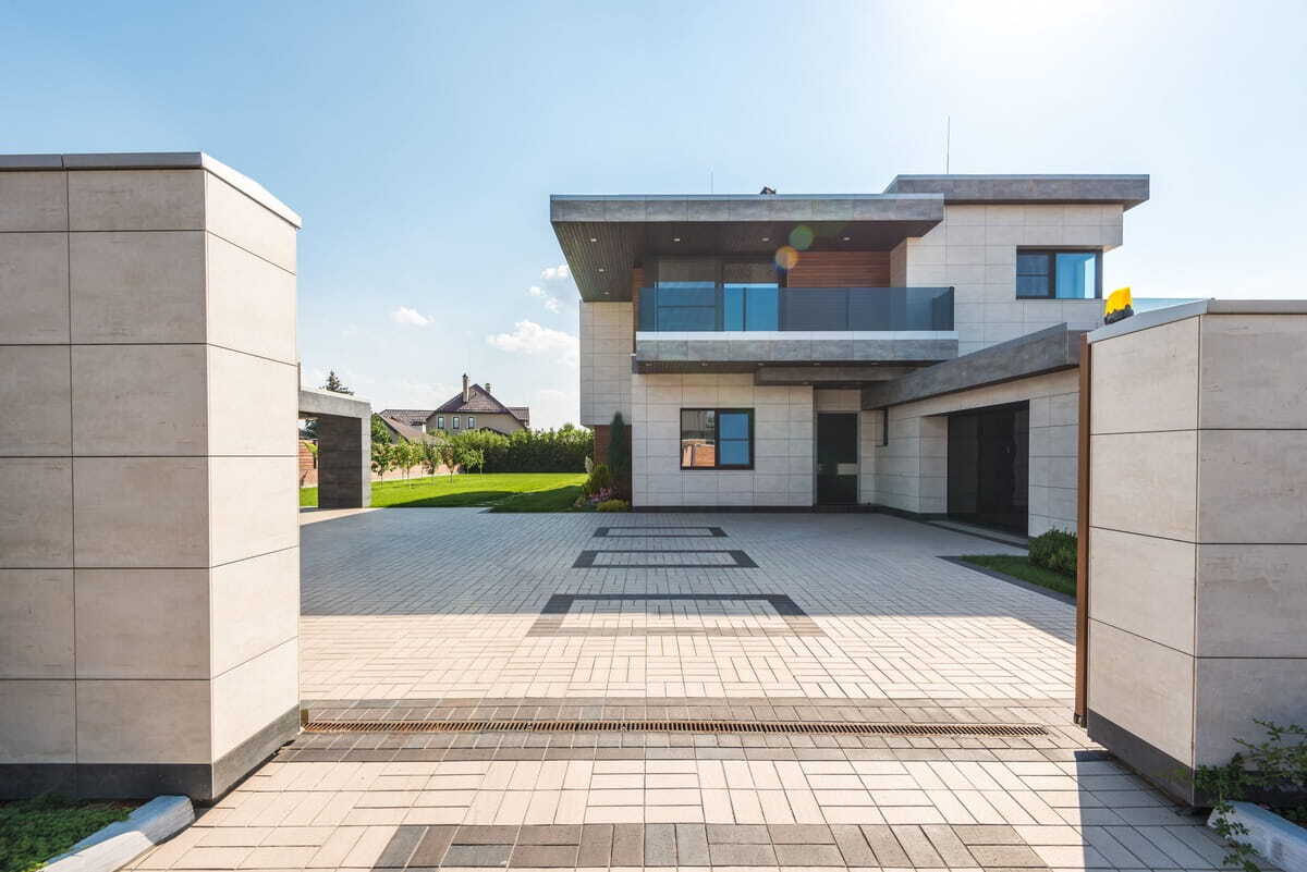 Modern two-story luxury home with light beige tile siding, glass balcony, and a large paved block driveway under a clear blue sky.