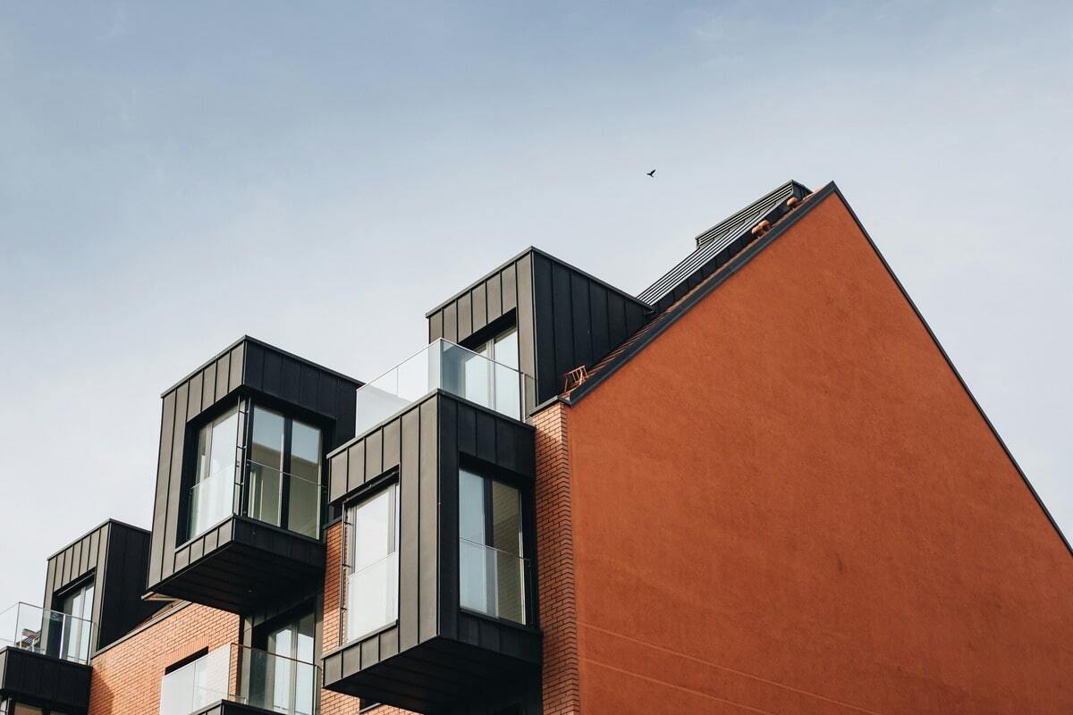 A modern building with a distinctive design, featuring a large, smooth, reddish-brown wall on the right and a section on the left with multiple levels of dark-colored, box-like protrusions. These protrusions have large windows and glass balconies. The building is made of brick and what appears to be dark metal cladding, symbolising the decent homes standard. A clear sky is visible in the background, with a single bird flying.