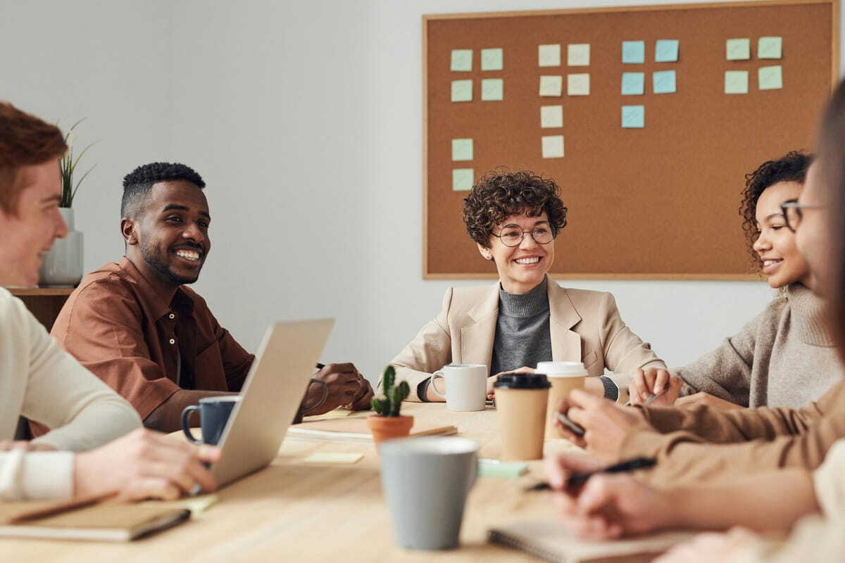 A diverse team of professionals collaborates around a wooden table in a bright office, with coffee and a sticky-note brainstorm board in the background.