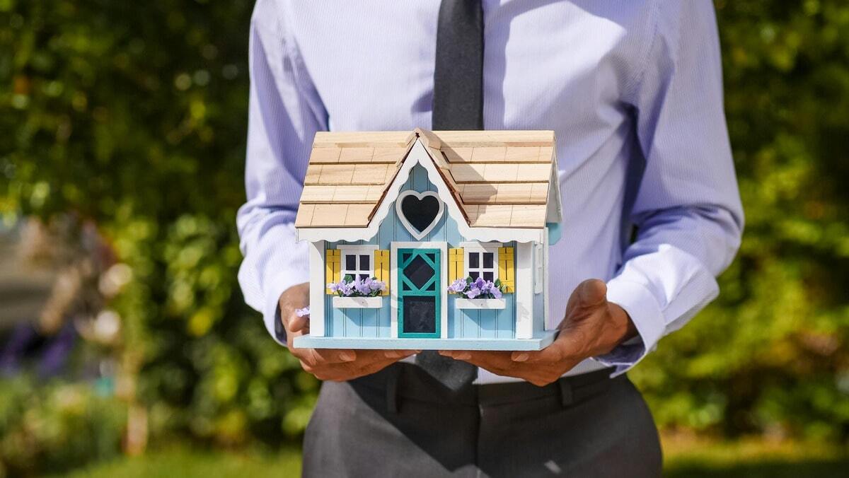 A man in a blue dress shirt and tie holds a small, mint-green wooden model house with a brown roof, a heart-shaped window, and yellow shutters, standing in a sunny, green outdoor setting.