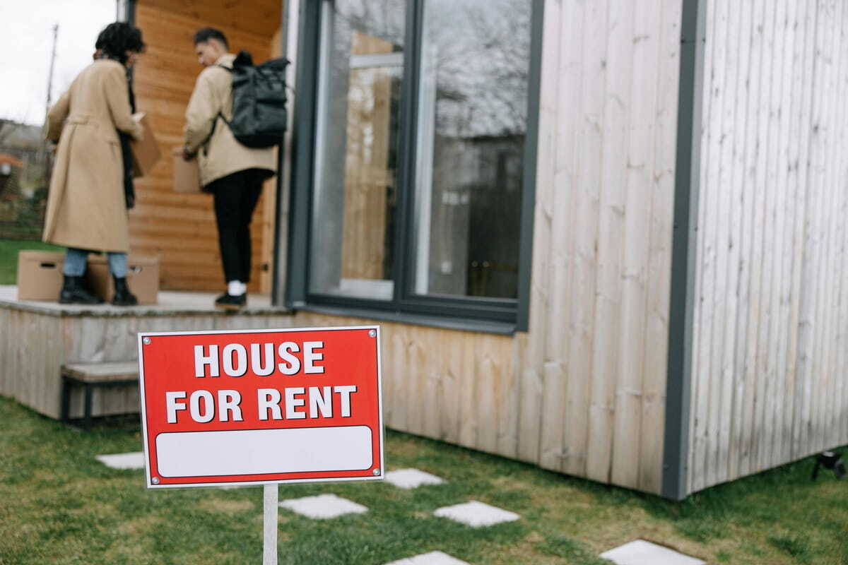 A red "House For Rent" sign stands in the foreground of a grassy yard. In the blurred background, two people carrying cardboard boxes walk toward the porch of a modern, light-wood-sided tiny house. One person wears a tan coat and jeans, while the other wears a tan jacket and a black backpack.