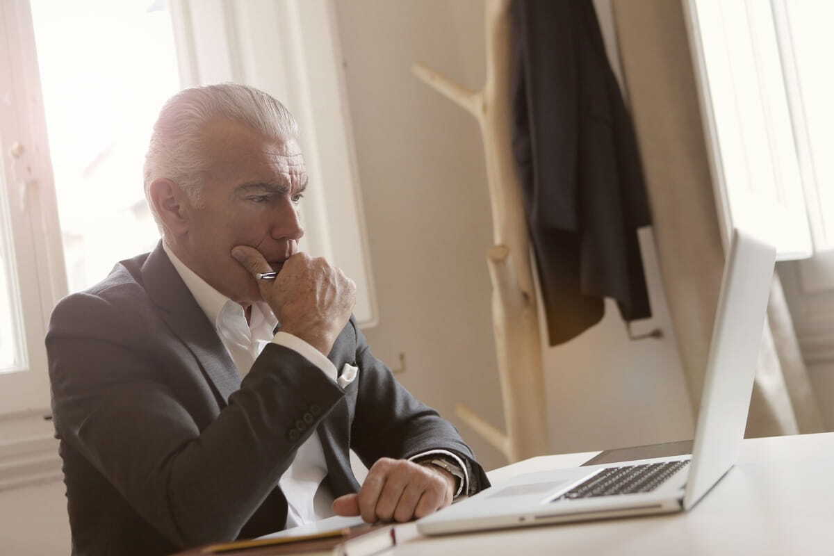 A senior businessman in a suit sitting at a desk, thoughtfully looking at his laptop screen.