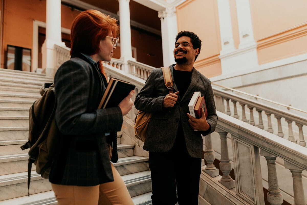 Two smiling students, a woman with red hair and a man with dark curly hair, holding books and wearing backpacks while chatting on a grand marble staircase.