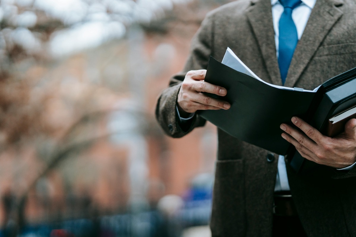 Close-up of a person in a brown tweed blazer, white shirt, and blue tie, holding open a black folder with documents outdoors.