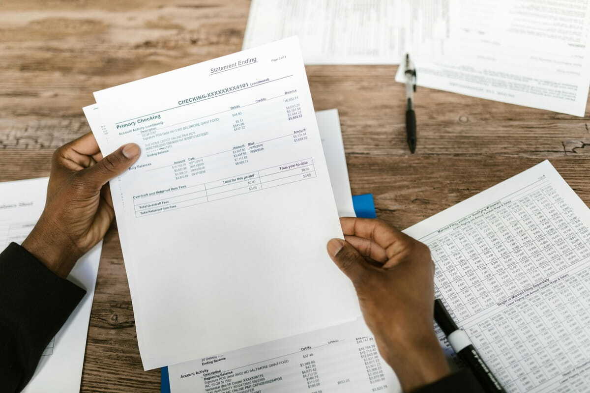 A top-down view of a person with dark skin holding a printed bank statement over a wooden desk scattered with other financial documents and a black pen. The statement in focus shows account activity for a "Primary Checking" account, including columns for descriptions, debits, credits, and balances. To the right, another document displaying a complex tax or financial table is visible.