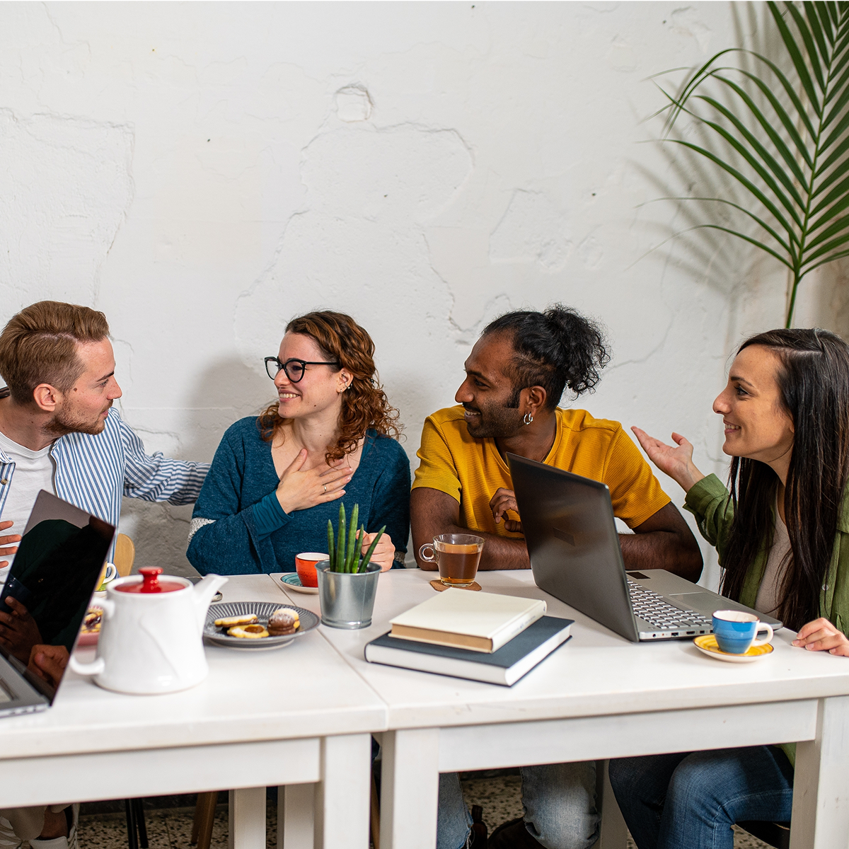 Group of people sitting at a table in an office, talking and smiling while working on laptops and sharing drinks.