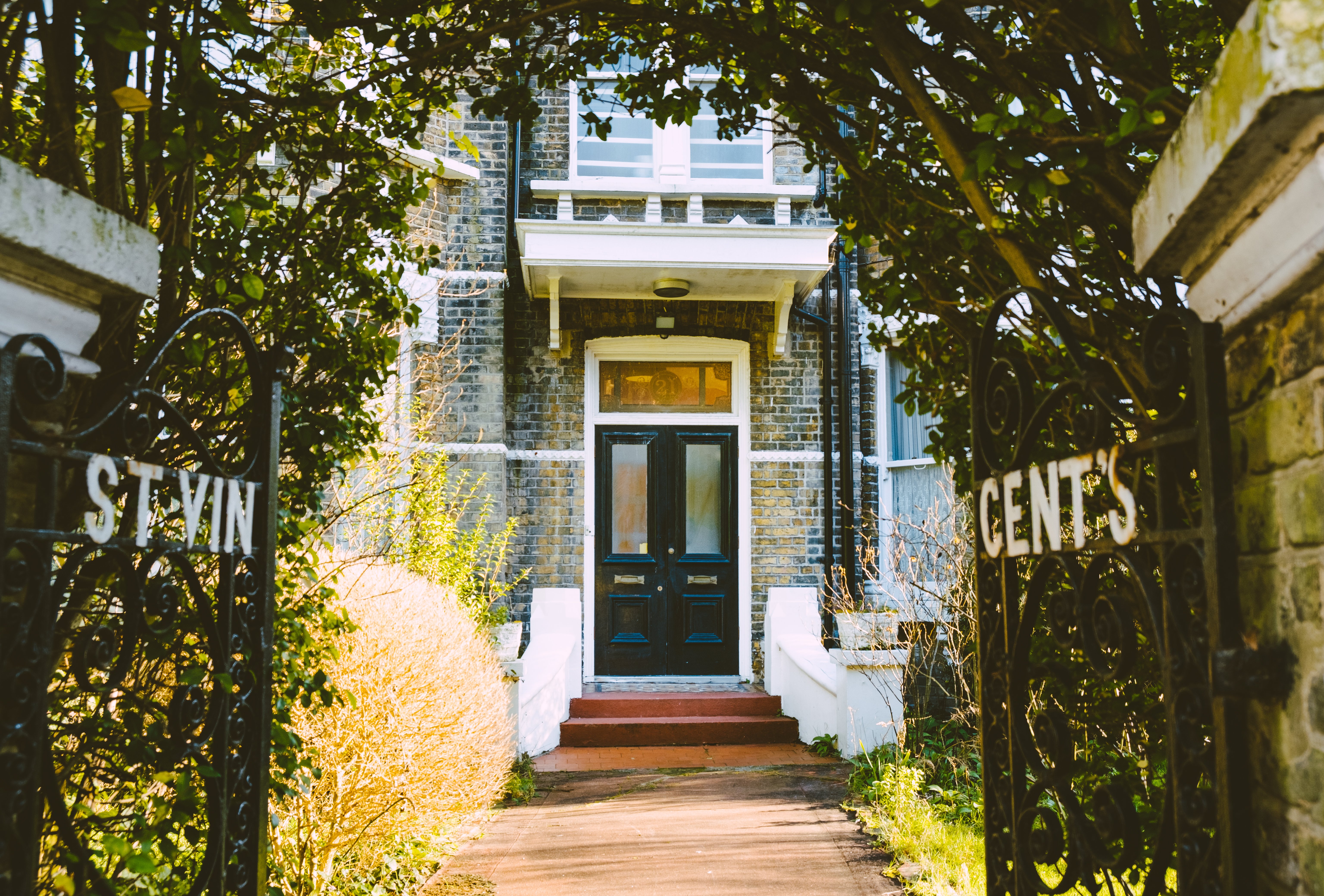 Open black gates with the text "St Vincent's" in white paint, followed by a brown garden path to a black door with grass and plants on either side. 