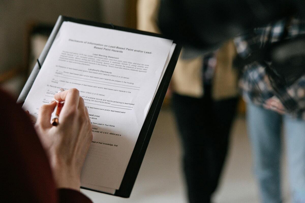 A close-up shot of a person's hands holding a pen and filling out a form on a clipboard, which appears to be a disclosure of information. In the blurred background, another person is standing.