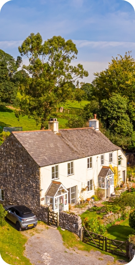 A two-story stone and cream-colored cottage with two small enclosed porches sits nestled on a green hillside with mature trees. A modern black car is parked beside the stone driveway.