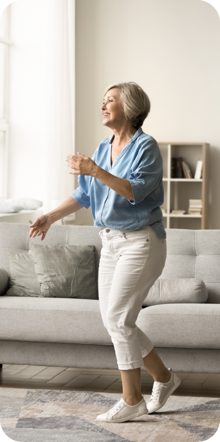 A happy, smiling older woman with short gray hair dances energetically in her living room, wearing a light blue shirt and white pants.
