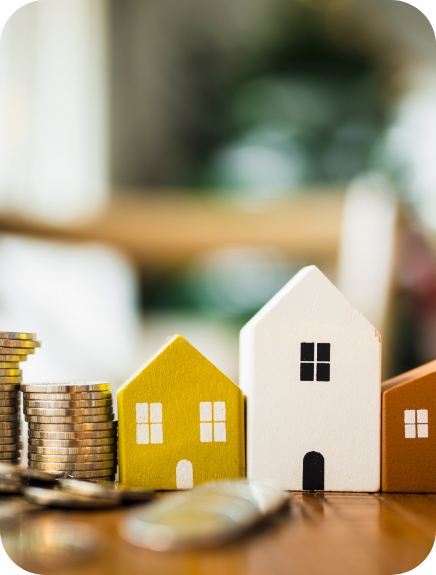 Stacked coins beside small wooden model houses on a table, with a blurred background.