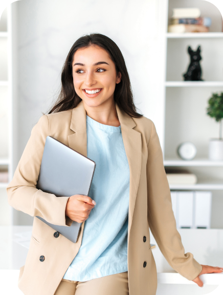Smiling woman in a beige blazer holding a laptop, standing in a bright office setting with shelves in the background.