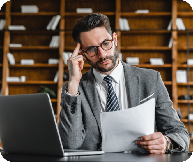 Man in a suit and glasses sitting at a desk with a laptop, holding and reading a document with a thoughtful expression, with bookshelves in the background.