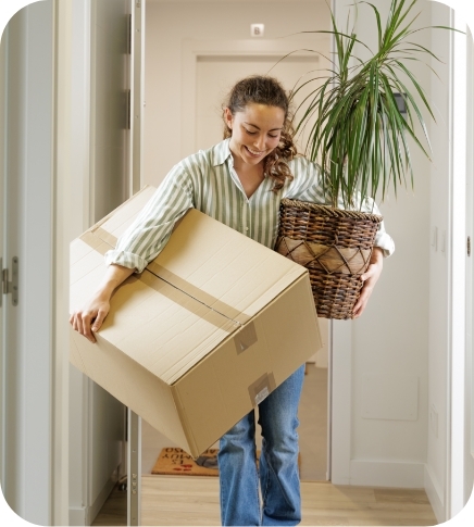 A smiling young woman in a striped shirt and jeans carries a large cardboard moving box and a potted dracaena plant down a hallway, representing moving into a new home.