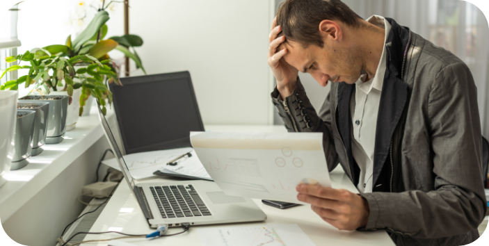 Man sitting at a desk with a laptop and documents, holding his head in frustration while reviewing paperwork.