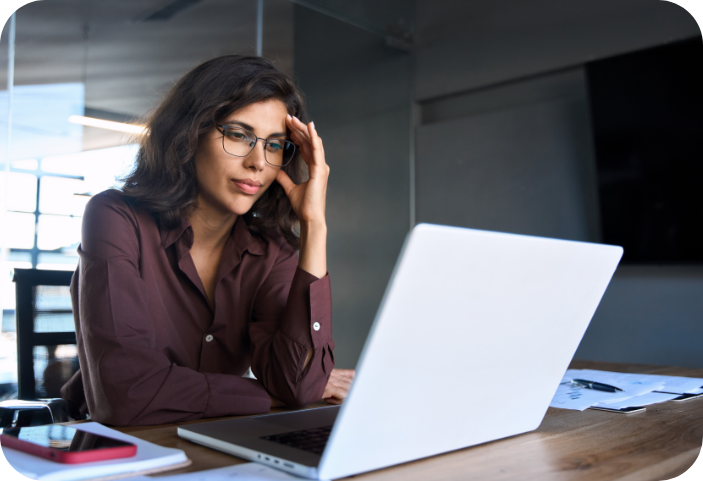 Woman wearing glasses sitting at a desk with a laptop, resting her head on her hand while concentrating on the screen.