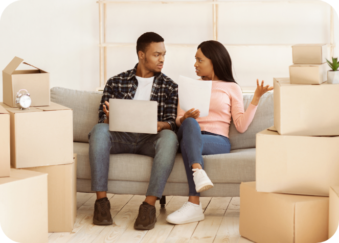 Couple sitting on a couch surrounded by moving boxes, looking concerned while discussing paperwork and using a laptop.