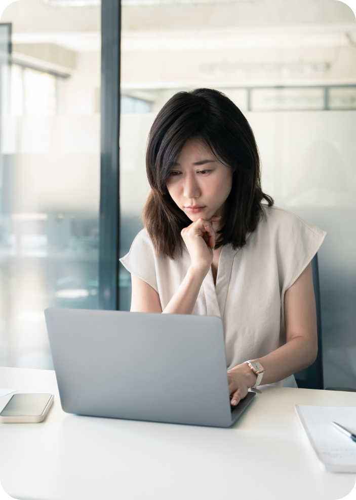 Woman sitting at a desk working on a laptop, resting her chin on her hand with a thoughtful expression in an office setting.